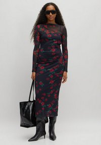 Enganliegendes, langärmliges Kleid aus durchscheinendem schwarzem Stoff mit einem floralen Rosenmuster in Rot und Grün. Kombiniert mit schwarzen knielangen Stöckelschuhen und einer Handtasche.