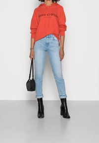 Felpa rossa con logo, jeans slim-fit azzurri con orli risvoltati, stivali neri con tacco e una piccola borsa nera. Tessuti con texture lisce.