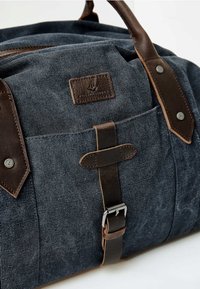 Torba podróżna z materiału płóciennego w ciemnoniebieskim kolorze z akcentami ze brązowej skóry. Posiada przednią kieszeń z klapką i detalem w postaci zapięcia, wzmocnione uchwyty oraz fakturowane wykończenie.