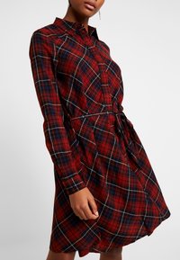 Punainen ruudullinen mekko pitkillä hihoilla, napitusyksityiskohdilla, vyötäröllä solmittavalla vyöllä ja kaarevalla helmalla. Valmistettu pehmeästä, kevyestä kankaasta.