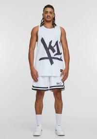 White mesh sleeveless tank top with black graphic lettering, paired with white shorts featuring black stripes and accents. White sneakers and socks.