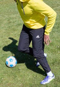 Žlutá mikina s kapucí, černé sportovní kalhoty, bílé ponožky a fialové kopačky na fotbal. Trávník s modrobílým fotbalovým míčem. Dynamická pozice.