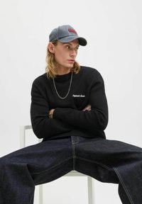 Black sweatshirt with white text, oversized dark denim jeans with visible stitching, gray cap with red lettering, long blonde hair, neutral background.