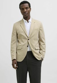 Beige blazer with a textured fabric, single-button closure, two front pockets, worn over a white collared shirt, paired with dark trousers.