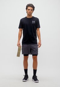 Νέος άνδρας που στέκεται φορώντας αθλητική ενδυμασία σε μαύρο και γκρι, κρατώντας μια μπεζ λωρίδα αντίστασης, με απλό ανοιχτό γκρι φόντο.