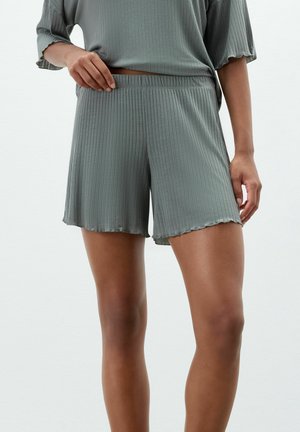 Vrouw draagt geribde, losse groene shorts en bijpassende top, houdt met één hand de tailleband van de shorts vast.