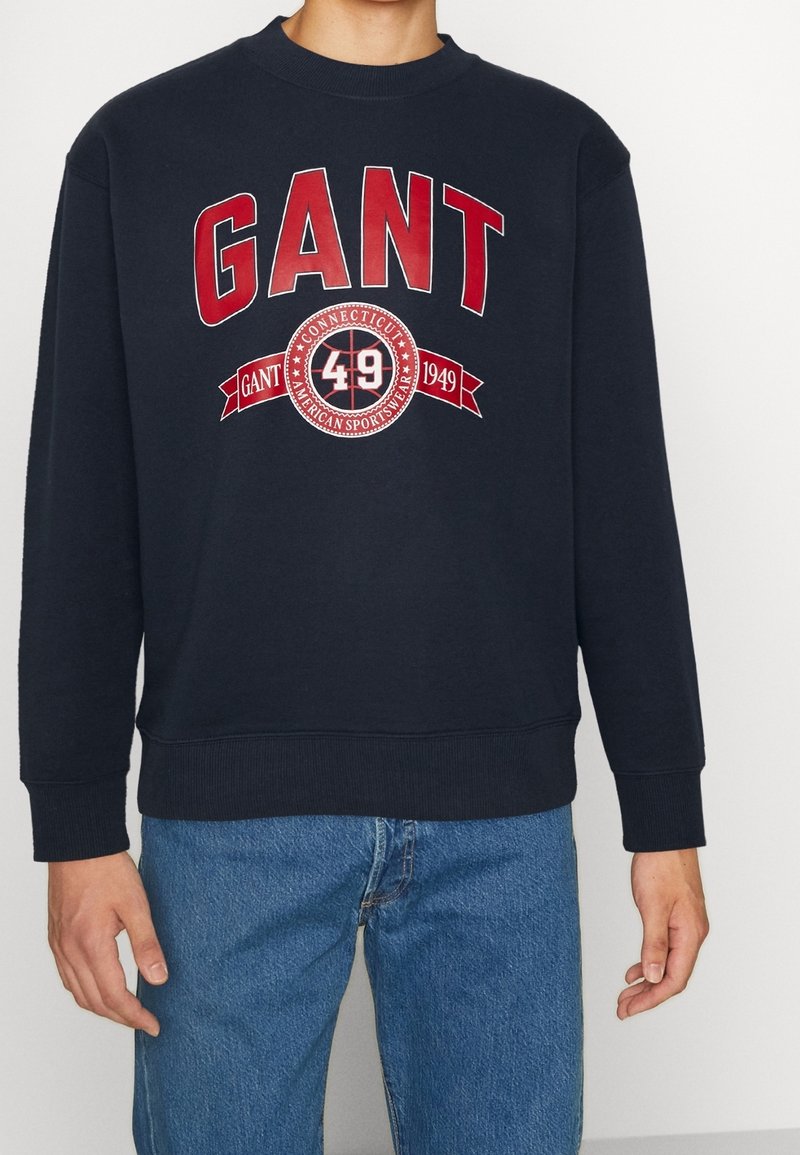Sudadera de marina con grandes letras rojas "GANT", logotipo circular con "49" y "1949", puños de canalé y cuello redondo. Ajuste casual.