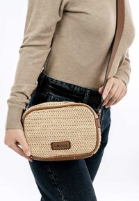 Bolso cruzado de paja trenzada con detalles en marrón, cierre con cremallera, forma cuadrada y correa ajustable. Superficie texturada con detalle de logo.
