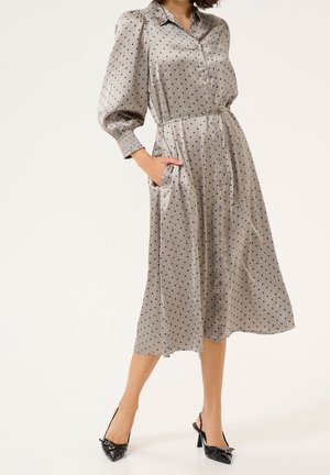 Kvinde iført en grå satin midi-kjole med sorte polkaprikker, puffede ærmer, knapper foran og sorte højhælede sko.
