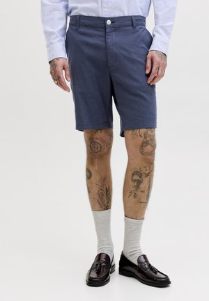 Άνδρας που φοράει σκούρα μπλε σορτς, ανοιχτό μπλε ριγέ πουκάμισο, ανοιχτό γκρι κάλτσες, σκούρα καφέ loafers με φούντες, με ορατά τατουάζ στο πόδι και το χέρι.