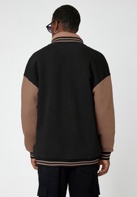 Sudadera color bloqueada en negro y marrón con cuello alto, puños y parte inferior acanalados. Textura suave con mangas y rayas en contraste.