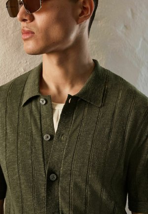 Man wearing dark green buttoned knit shirt and sunglasses, shown from side and chest up against textured wall background.
