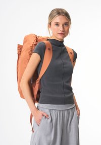 Oranger Stoffrucksack mit verstellbaren Trägern, Reißverschlusstaschen und Rollverschluss. Das Model trägt ein dunkles Oberteil und einen grauen Rock, die Hände in den Taschen.