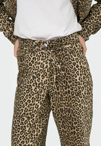 Los pantalones de leopardo presentan un fondo beige con manchas marrón oscuro, un corte relajado, trabillas para cinturón y un detalle de lazo en la parte delantera.