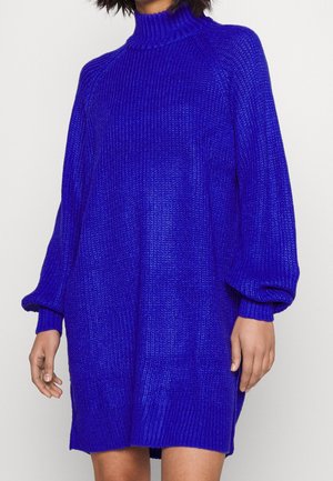 Femme portant une robe-pull tricotée ample, à manches longues, de couleur bleu vif, avec un col haut et des poignets côtelés.