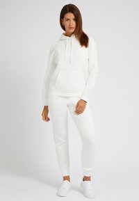 Vit bomullshoodie med framficka och dragsko, tillsammans med matchande slim-fit joggers. Slät textur och ribbade ärmslut. Vita sneakers.