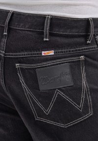 Juodi džinsai su kontrastingais baltais siūlais, odiniu etikete su įspaustu "Wrangler" ir "W" logotipu ant nugarinės kišenės.