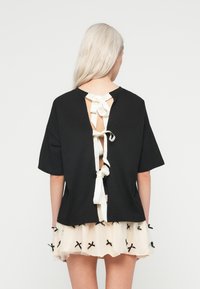 Pessoa com cabelo loiro comprido, vestindo uma blusa preta de mangas curtas com três laços brancos na parte de trás e uma saia bege com laços pretos.