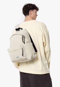 Béžový kordurový batoh se dvěma zipovými kapsami, nastavitelnými popruhy a logem, nošený na zádech osoby v bledém topu.