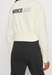 Sweatshirt crème coupé avec "NIKE AIR" brodé en maille noire dans le dos ; associé à un pantalon gris foncé, affichant une coupe détendue.