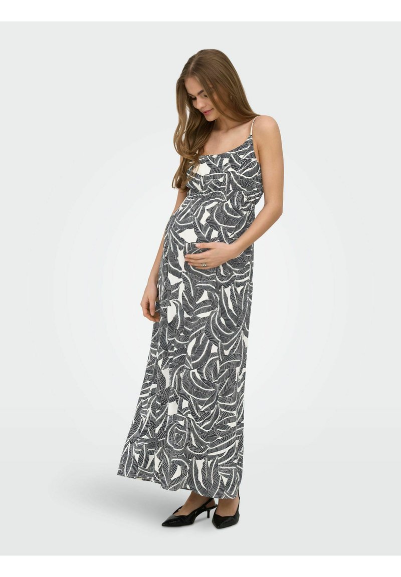 Femme enceinte en robe maxi sans manches à motif de feuilles noir et blanc, tenant doucement son ventre, portant des talons noirs, debout sur fond blanc.