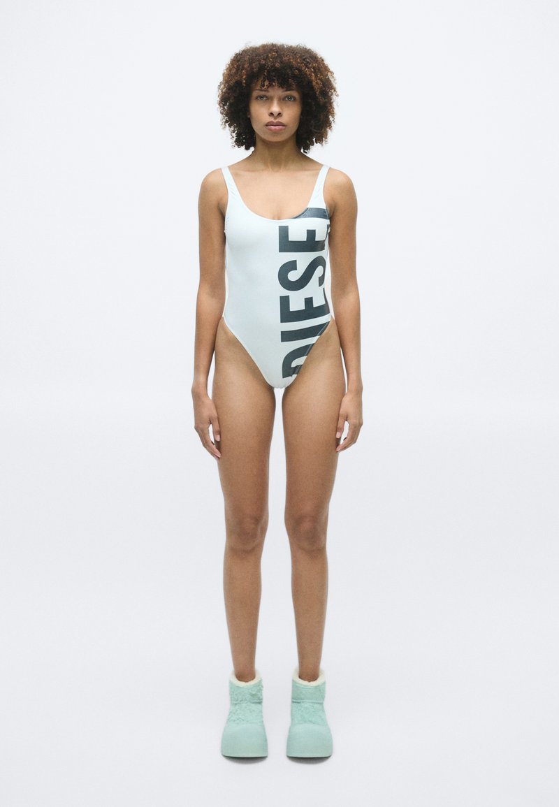 Femme aux cheveux bouclés portant un maillot de bain une pièce bleu clair avec le texte "DIESEL" et des pantoufles moelleuses vert menthe, se tenant sur un fond blanc.