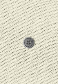 Tessuto a maglia color panna con motivo a rilievo; presenta un bottone metallico rotondo inciso con la scritta "THE ORIGINAL."