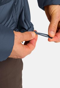 Eine blaue isolierte Jacke wird an der Taille mit einem Kordelzug angepasst. Die Hände greifen das elastische Band zum Festziehen, wodurch die Textur des Stoffes sichtbar wird.