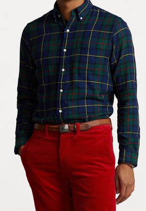 Mann trägt ein grün-blau kariertes Hemd mit Knöpfen, das in rote Cordhosen mit braunem Ledergürtel gesteckt ist.