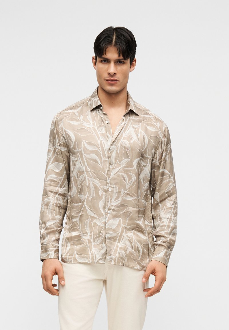 Homme portant une chemise beige à manches longues avec motif de feuilles blanches et un pantalon blanc cassé, debout devant un fond clair uni.