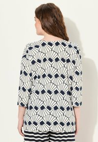 Femme aux cheveux bruns ondulés de dos, portant un haut crème et noir à motif en éventail et un pantalon rayé noir et blanc.