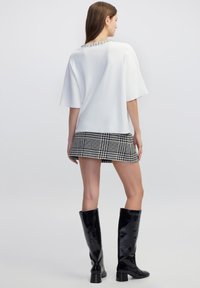 Top blanco de manga corta con escote decorado, combinado con una falda corta de cuadros en blanco y negro. El modelo lleva botas negras brillantes hasta la rodilla.