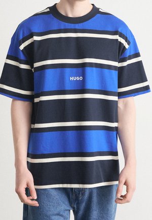 Mann trägt blaues, schwarzes und weißes T-Shirt mit horizontalen Streifen und dem Schriftzug „HUGO“ auf der Brust, kombiniert mit blauen Jeans, Oberkörper sichtbar.