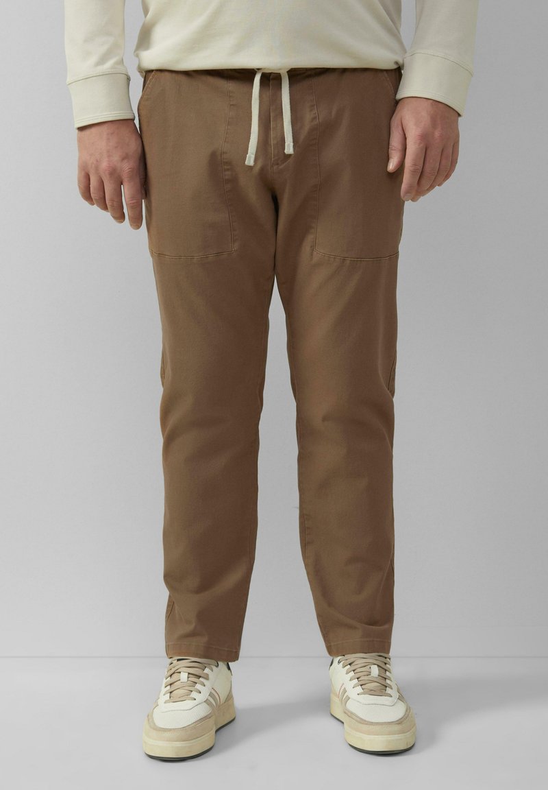 Bruine katoenen cargobroek met voorzakken, een trekkoord tailleband en taps toelopende pijpen, gecombineerd met witte en beige sneakers.