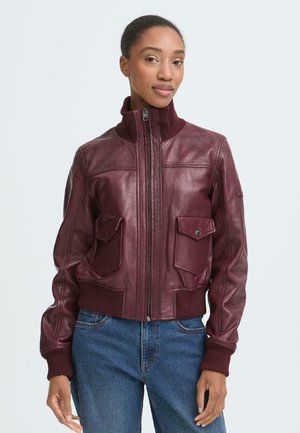 Donna che indossa una giacca in pelle bordeaux con cerniera, polsini a coste, colletto alto, tasche con patta e jeans blu su uno sfondo semplice.