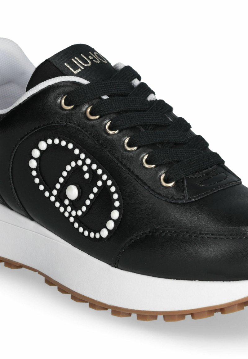 Sneaker en cuir noir avec des détails en accent blanc, présentant un design à lacets, un bout rond et une semelle épaisse blanche avec un fond texturé.