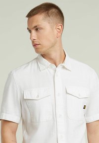 Camisa blanca de manga corta hecha de tejido ligero, con dos bolsillos en el pecho, cierre de botones y una etiqueta de marca negra en el lateral.