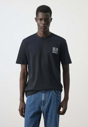 Joven con una camiseta negra con un pequeño parche en el pecho y vaqueros azules frente a un fondo claro y liso.