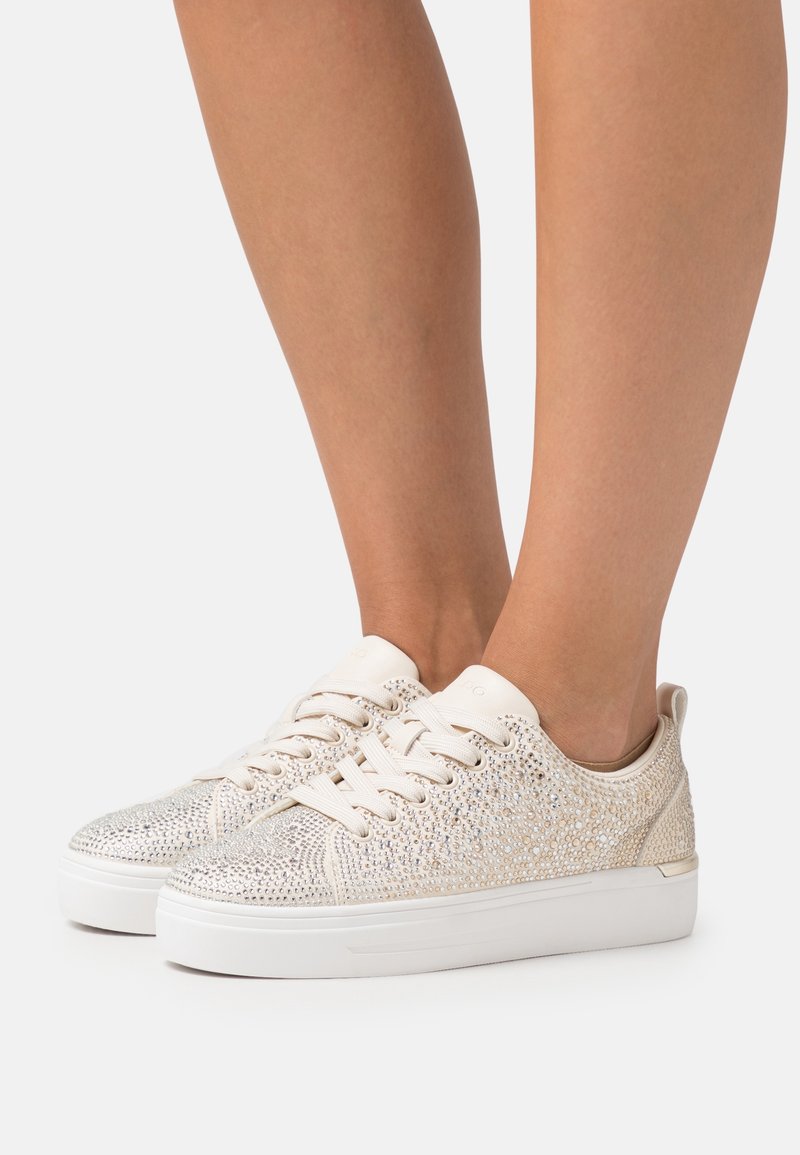 Sneakers with a beige shimmer mesh upper, covered in silver rhinestones, and a white rubber sole. Laced design with cushioned heel.