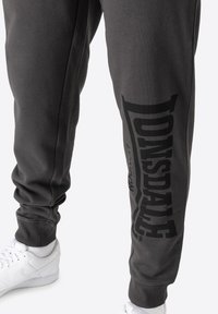 Grå sweatpants lavet af en blanding af bomuld, med et stort sort logo trykt på venstre ben og ribbede manchetter, parret med hvide sneakers.