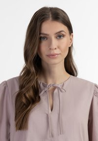 Lyse mauve bluse med rund halsudskæring, nøglehulåbning og bundet bånddetalje. Har pufærmer og glat, tekstureret stof.