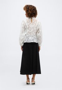 Γυναίκα με σγουρά μαλλιά φορώντας λευκή δαντελένια μπλούζα με μακριά μανίκια και μαύρη midi φούστα, στέκεται γυρισμένη με την πλάτη προς το μέρος μας σε απλό φόντο.