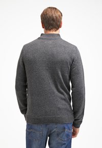 Suéter de punto gris oscuro con un cuello alto, puños y dobladillo acanalados, y una superficie texturizada. Se lleva con jeans de denim azul casuales.