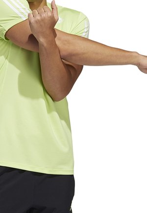 Personne portant un t-shirt Adidas vert citron étirant le bras droit à travers la poitrine avec la main gauche.