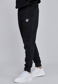 Czarne dresy z białym logo na lewym udzie, bocznymi kieszeniami i ściągaczami na nogawkach. W zestawie z białymi sneakerami na szarym tle.