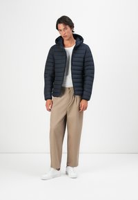 Jonge man in een zwart gewatteerd jack met capuchon, een wit T-shirt, beige wijde broek en witte sneakers, staande tegen een witte achtergrond.