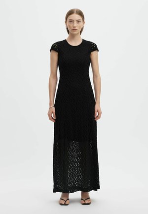 Femme portant une robe longue noire en dentelle à manches courtes avec une doublure unie arrivant à mi-cuisse, associée à des sandales noires à bouts ouverts.