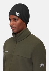 Zwarte gestreepte muts met een wit olifantenlogo, gedragen door een persoon in een groene fleecejack met rits en logo-detail op de mouw.