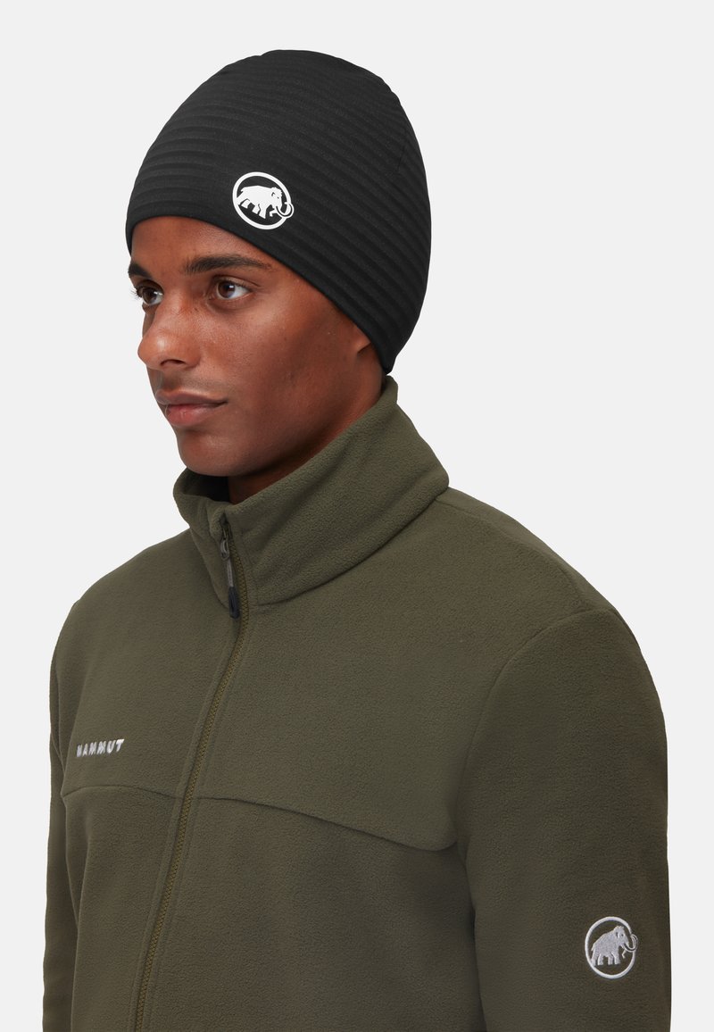 Zwarte gestreepte muts met een wit olifantenlogo, gedragen door een persoon in een groene fleecejack met rits en logo-detail op de mouw.