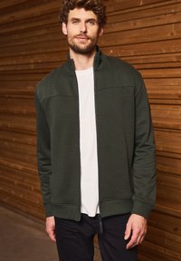 Veste zippée pour homme en vert foncé avec un motif hexagonal texturé sur le haut du corps. Dispose de poignets côtelés et d'un t-shirt blanc uni en dessous.